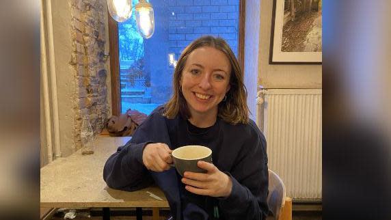 Gemma is sitting at the table holding a mug and smiling, wearing dark clothes.