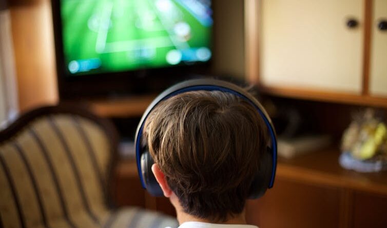 Back view of a boy wearing headphones and looking at a TV screen.