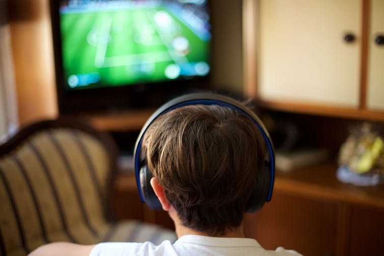 Back view of a boy wearing headphones and looking at a TV screen.