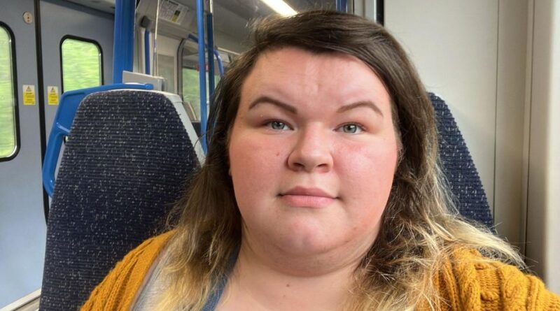 Nikki, who has brown hair and is wearing a yellow knitted cardigan, is sitting in a train carriage with an empty seat next to her.  He looks directly at the camera, his face neutral.